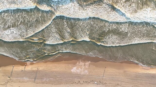 Aerial top down view of powerful ocean waves crashing on a sandy beach. Scenic drone perspective of sea surf and coastal nature. Natural texture of water foam and shoreline.