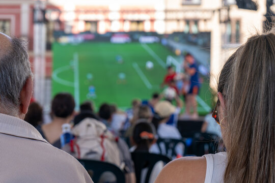 People watching football match on large outdoor screen before game start at public viewing event