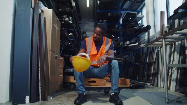 Tired African American warehouse worker taking off a yellow hard hat and sitting down to rest after a hard working day.