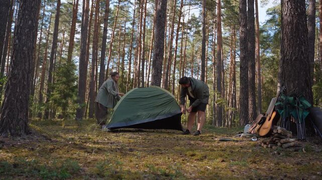 Young man and woman pitching a green tourist tent together in a sunny pine forest, preparing their campsite for an outdoor adventure and enjoying an active weekend getaway in nature
