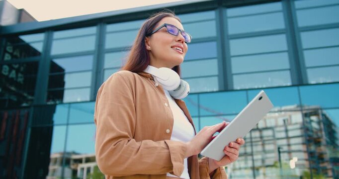 Side view of a young woman student holding a tablet outdoors, standing on a university campus and browsing online study resources