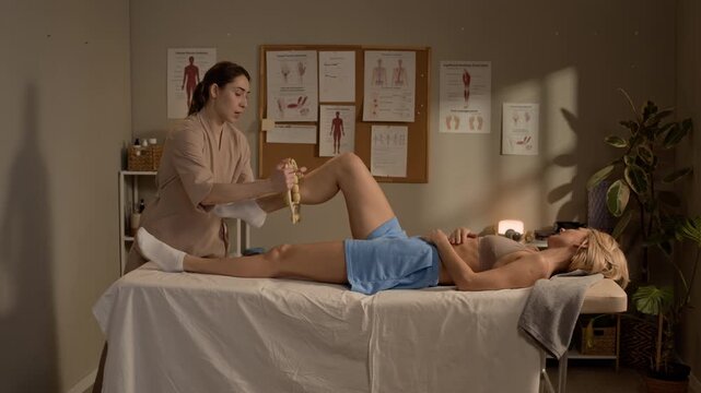 Wide shot of female massage therapist using wooden massage tool on blonde female client knee during wellness treatment in cozy therapy room