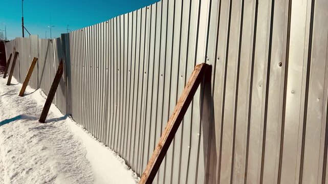 Snow-covered corrugated steel fence leaning on wooden braces, winter construction site under bright blue sky, deep snowdrifts piled