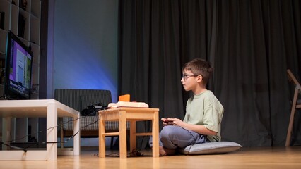 Young boy wearing glasses sitting on the floor in a dark room, concentrically playing old school video games on a classic console connected to a modern television at home © Aliaksandr
