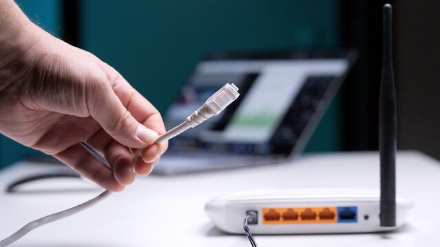 Person's hand holding and connecting an ethernet cable to a white wireless router, providing a wired internet connection while a laptop with a screen is visible in the blurred background