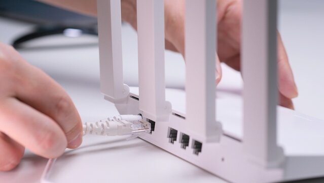 Person hand connecting a white ethernet cable into the port of a modern white wireless router with multiple external antennas, establishing a network connection for internet access