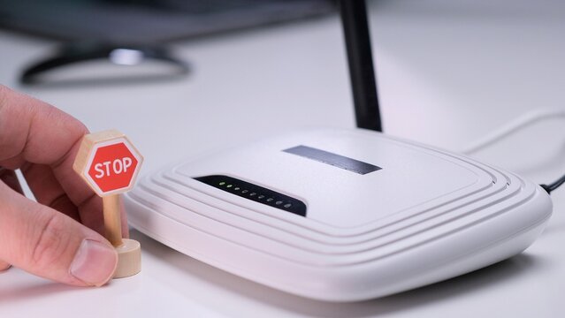 Person placing a small red stop sign in front of a white wireless router with illuminated indicator lights, illustrating internet access restriction, control, or interruption