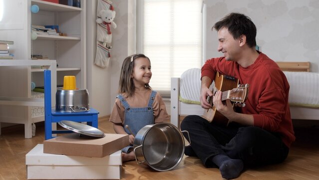 Father and daughter are enjoying a creative and joyful moment together, bonding through music as the dad plays guitar while the child smiles next to her homemade pot and pan drum set at home