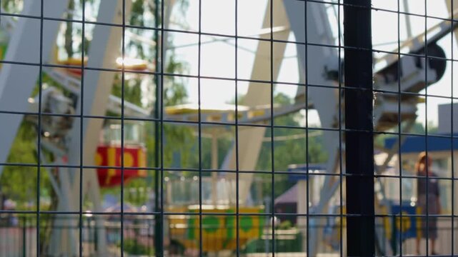 Metal grid in foreground creates patterned overlay, partially obscuring vibrant Ferris wheel in background. The contrast between rigid lines of fence and playful, colorful cabins evokes sense of antic