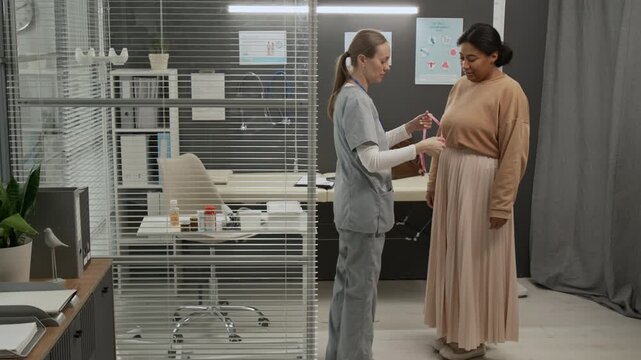 Female medical specialist in scrubs taking sewing tape from her pocket and measuring waist of woman during fertility consultation