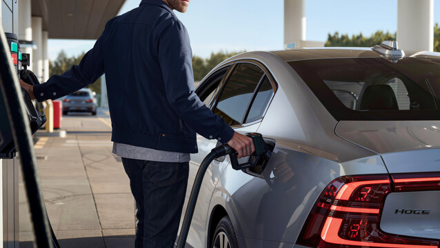 ​Man refuels silver sedan at station.
​Rear view of car's red taillights.
​Driver standing beside fuel pump station.
​Filling up vehicle for road trip.
​Modern sedan during early morning refueling.
