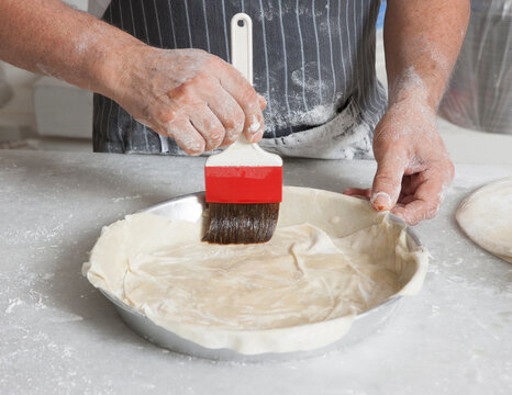Hands brushing phyllo dough with oil in a baking tray. Traditional homemade pie preparation process in kitchen, ready for baking.
