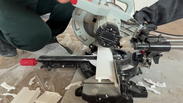 Worker cutting duropolymer skirting board using miter saw on floor during apartment renovation. Close-up of hands and tools, construction process, interior finishing work.