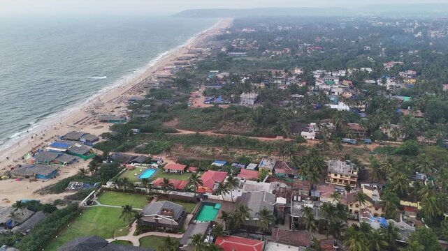Cinematic aerial view of a sprawling coastal town where dense palm-covered neighborhoods meet a long sandy shoreline, with waves rolling in along the edge of the city.