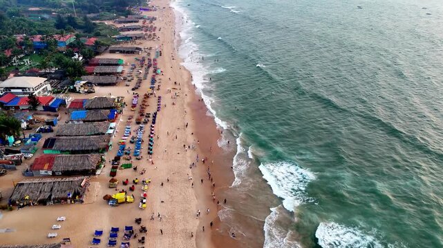 Wide close cinematic aerial of a bustling beach featuring patterned sunbeds, scattered beachgoers, and rolling waves. The blend of geometry and natural motion enhances the visual depth.