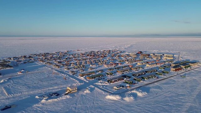 Aerial orbit over Arviat, Nunavut at golden hour sunset. Long shadows stretch across colorful houses and snow-covered streets on the flat Hudson Bay tundra.