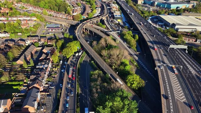 Drone flying along M6 motorway slip road near Birmingham, UK. Red bus, trucks and cars on curved elevated ramp beside residential streets and green trees.