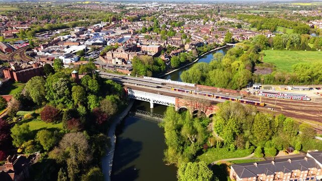 Drone approaching over River Severn as passenger train crosses Victorian railway bridge in Shrewsbury, UK. Red brick station, river loop and town panorama visible.