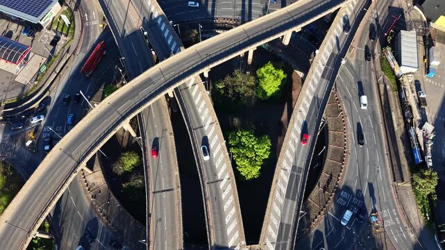 Straight down aerial drone shot of roundabout beneath elevated motorway flyovers in Birmingham, UK. Abstract geometric road pattern with green trees between concrete ramps.