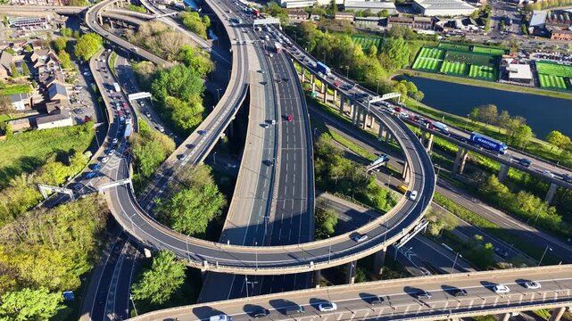 Drone flyover above Gravelly Hill Spaghetti Junction, Birmingham. Multi-level M6 ramps, heavy traffic, canal and green football pitches visible in background.