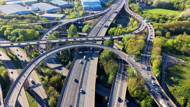 Drone flyover above Gravelly Hill Spaghetti Junction, Birmingham, UK. Multi-level M6 motorway interchange with flowing traffic, green trees and industrial buildings.