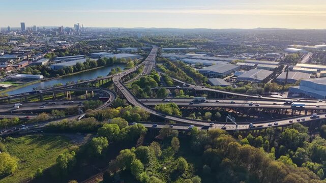 Static aerial drone shot of Spaghetti Junction M6, Birmingham. Traffic flows across multi-level ramps with river, green trees and city skyline in golden sunset light.