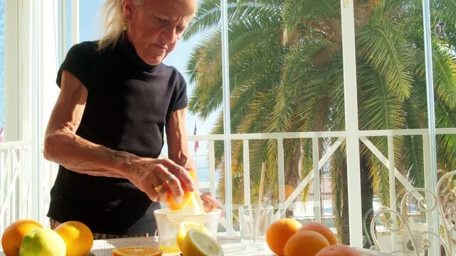 Senior woman squeezing fresh orange juice on a bright sunny terrace. Healthy lifestyle and Mediterranean breakfast concept with palm trees in the background during a peaceful morning.