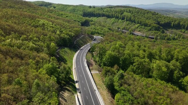 An aerial shot captures a newly built two-lane road curving through a dense forest, highlighting infrastructure's expansion into natural terrain.