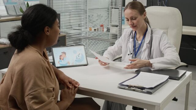 Black woman taking medical documents out of bag and giving them to Caucasian fertility doctor who then reviewing ultrasound results at white desk