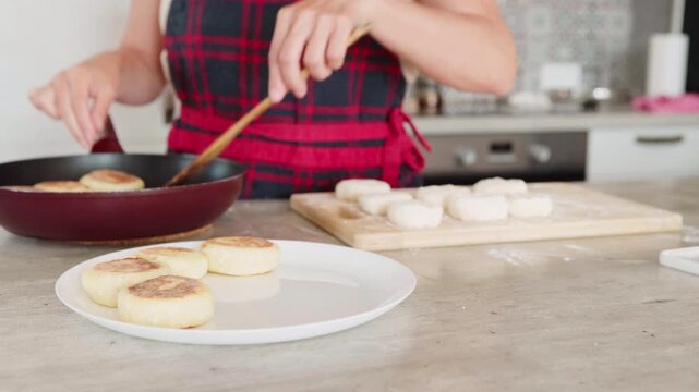 Close-up 4K footage of woman cooking syrniki fried Eastern Slavic quark (curd cheese) pancakes She places just-cooked meal on white plate in home kitchen. Healthy eating, breakfast preparation concept