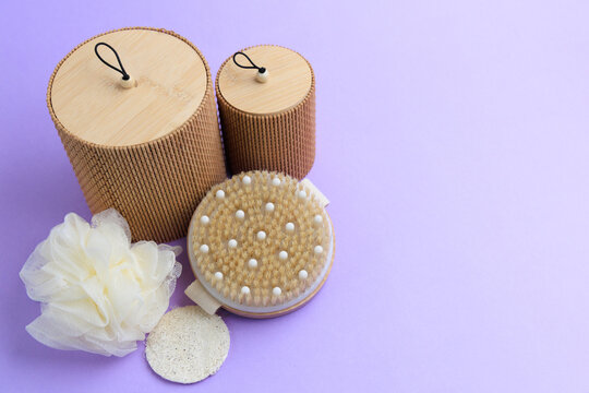 Collection of bath accessories on a light purple surface including loofah, scrub brush, and storage containers