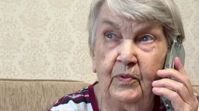 Elderly white woman holding smartphone sitting in cozy living room closeup, engaged in attentive phone conversation