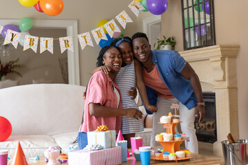 Naklejka premium African American family hugging daughter in living room, celebrating with cupcakes, gifts, balloons