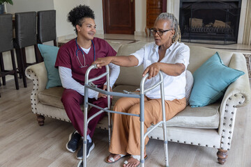 Naklejka premium Senior African American woman sitting on sofa at home using walker, caregiver in scrubs watching