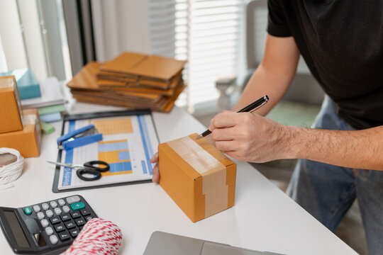 Small business owner writing on parcel box while preparing products for shipment, organizing orders and packaging items on desk, representing e commerce workflow and delivery process.