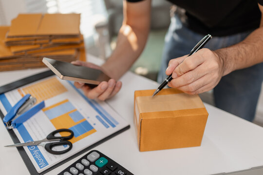 Small business owner checking order on smartphone while writing on parcel box, preparing packages for delivery, managing ecommerce workflow and shipping process on organized workspace.