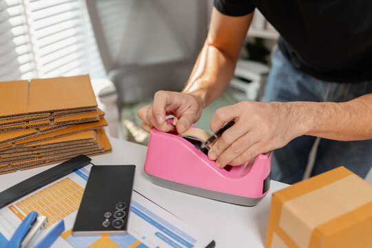 Small business owner sealing parcel with tape dispenser while preparing packages for shipment, organizing delivery orders and packaging process, representing ecommerce and logistics workflow.
