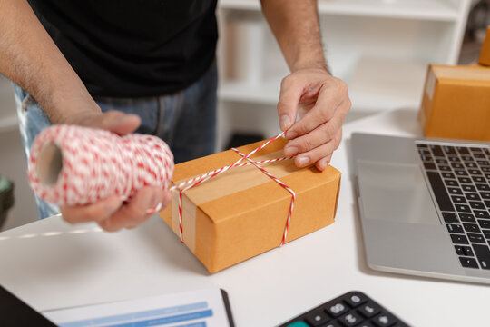 Small business owner tying parcel with string while preparing package for delivery, organizing shipping process and handling ecommerce orders, representing logistics and packaging workflow.