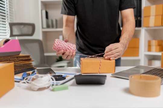 Small business owner tying parcel with string while preparing package for delivery, organizing shipping process and handling ecommerce orders, representing logistics and packaging workflow.