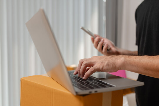 Small business owner typing on laptop while checking orders on smartphone, managing ecommerce sales and shipping tasks, representing digital business workflow and online order processing.
