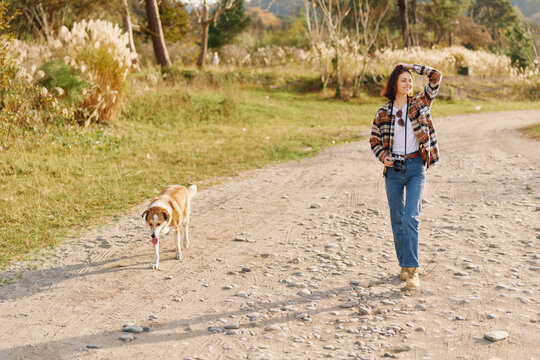 Woman walking with a dog along a dusty rural path, enjoying outdoors and nature, casual attire, camera ready as the sunlit countryside sets a peaceful mood