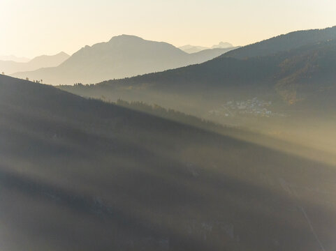 Mountain landscape with sun rays illuminating a misty forested valley and distant village, a serene tourism destination in Luserna, Alpe Cimbra, Trentino, Italy.