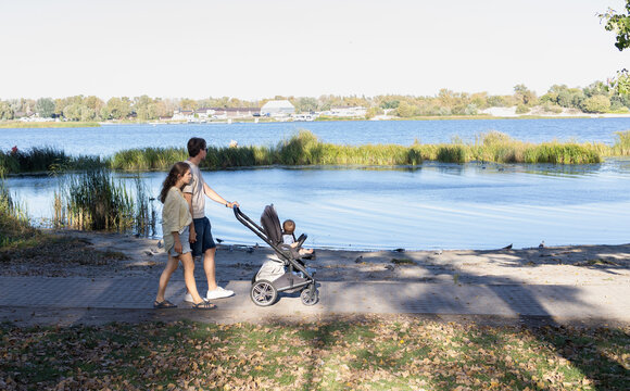 Young parents stroll with a stroller through a park next to a lake. The family is enjoying a walk together in nature on a summer day.