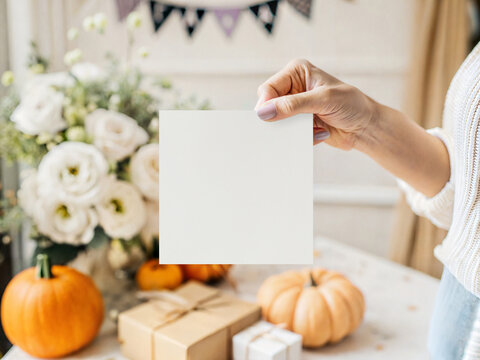 Aesthetic square 5x5 card mockup held by hand. Cozy autumn festive display with pumpkins, gifts and flowers on blurred interior background. Seasonal stationery branding template