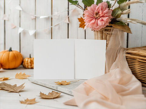 Aesthetic double square 5x5 card mockup with blank invitations on white wooden background. Cozy autumn display with basket of flowers, pumpkins and draped textile. Minimalist stationery branding