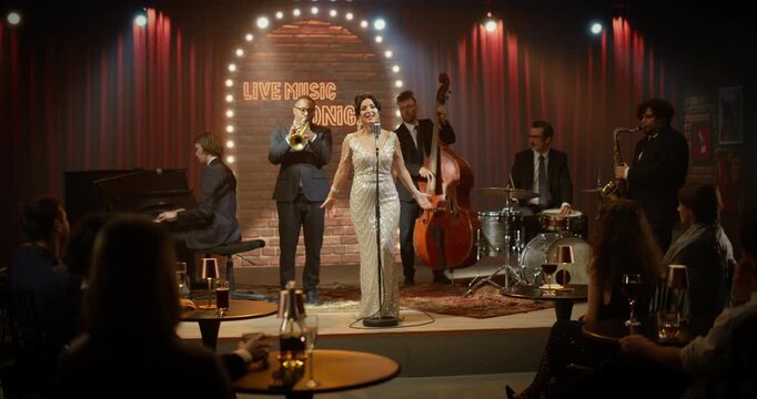 Stylish Female Singer in a Glittering Gown Sings at a Cozy Jazz Club Stage, Backed by Trumpet, Piano, Upright Bass, Drums and Saxophone as Guests Enjoy an Romantic Live Concert Night.