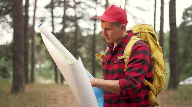 Hiker reading map in pine forest. Man with backpack studies map and looks around. Traveler navigates hiking trail in woods. Hiker checks route on paper map. Outdoor forest adventure with backpack.