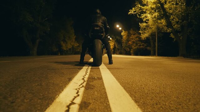 Biker in a helmet and leather jacket is sitting on his motorcycle at night highway and ready to ride. Man is going to ride a motorbike at evening time. Concept of freedom and adventure. Rear view