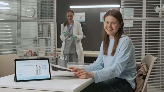 Portrait of cheerful Caucasian young woman with ultrasound images smiling at camera sitting at white doctors table, medical specialist examining stethoscope in background