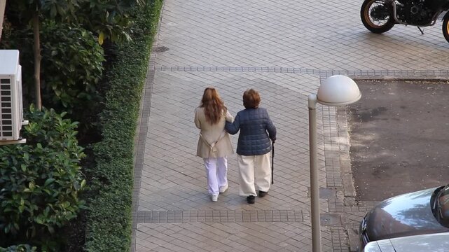 Anciana mayor dependiente con bast&oacute;n de apoyo y cuidadora asistente social del brazo de ayuda y compa&ntilde;&iacute;a como acompa&ntilde;ante en paseo por acerca de calle